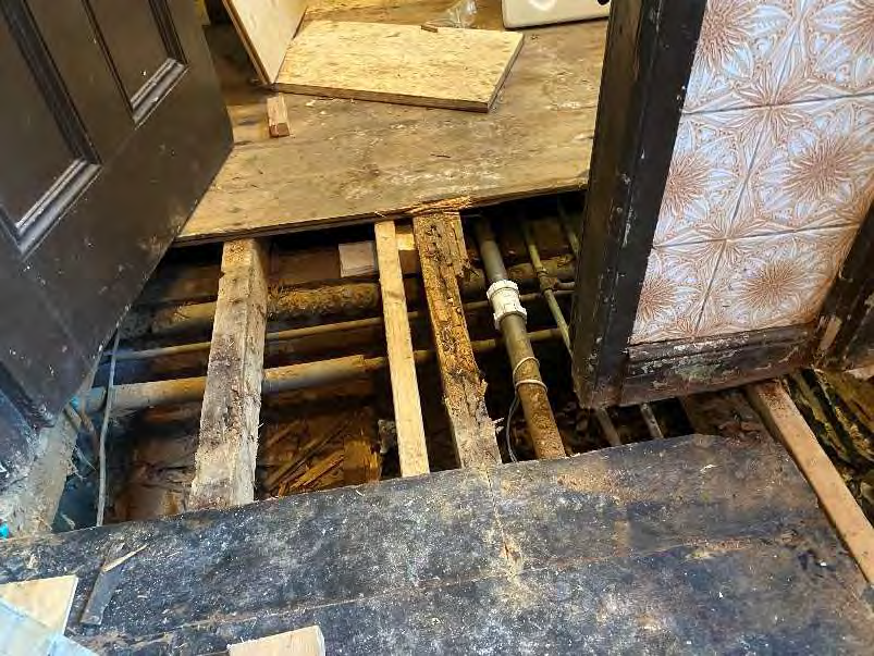A close-up photograph showing an interior floor under renovation with removed floorboards, exposing wooden joists and plumbing pipes underneath.