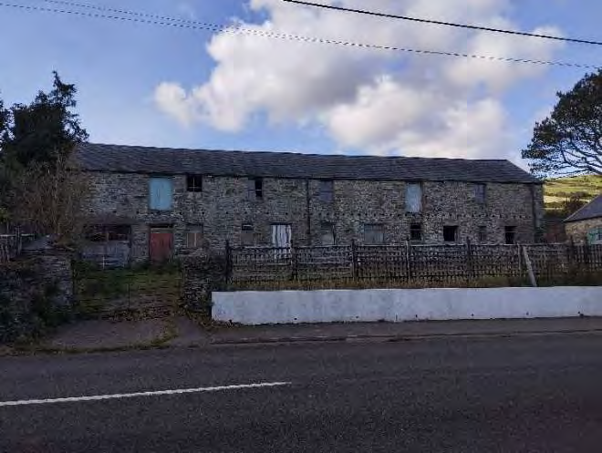 A photograph showing a long, single-story stone building with a slate roof, situated behind a low white wall and metal fence along a roadside.