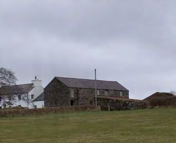 A photograph showing a rural site with a large stone building and an adjacent white house, set against a grassy foreground and overcast sky.