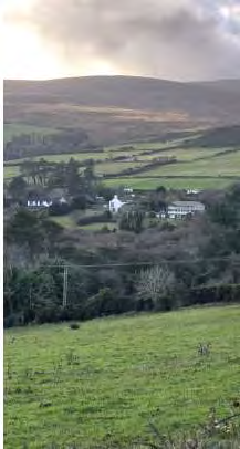 A scenic rural photograph showing a cluster of white houses nestled in a valley with green fields and rolling hills in the background.