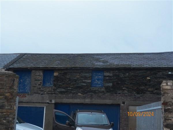 A photograph showing the exterior of a stone building with boarded windows and blue roller shutter doors, likely a barn or outbuilding.