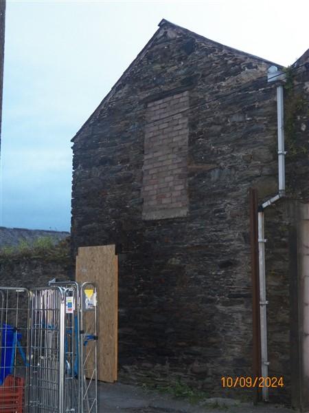 A photograph showing the side elevation of a stone building with boarded-up windows and doors, likely an outbuilding or barn, with a metal fence in the foreground.