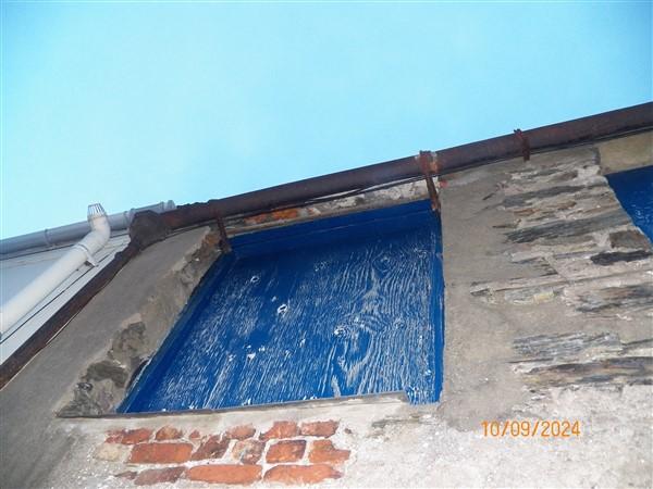 A low-angle photograph showing the upper wall and eaves of a stone building, featuring a blue painted boarded area, a rusty metal beam, and a white downpipe against a blue sky.