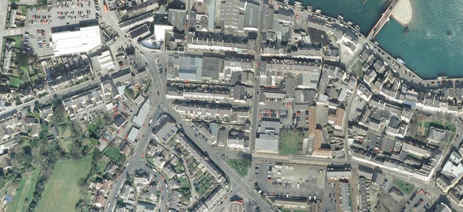 Aerial satellite view of a coastal town center showing streets, buildings, parking lots, and a harbor.
