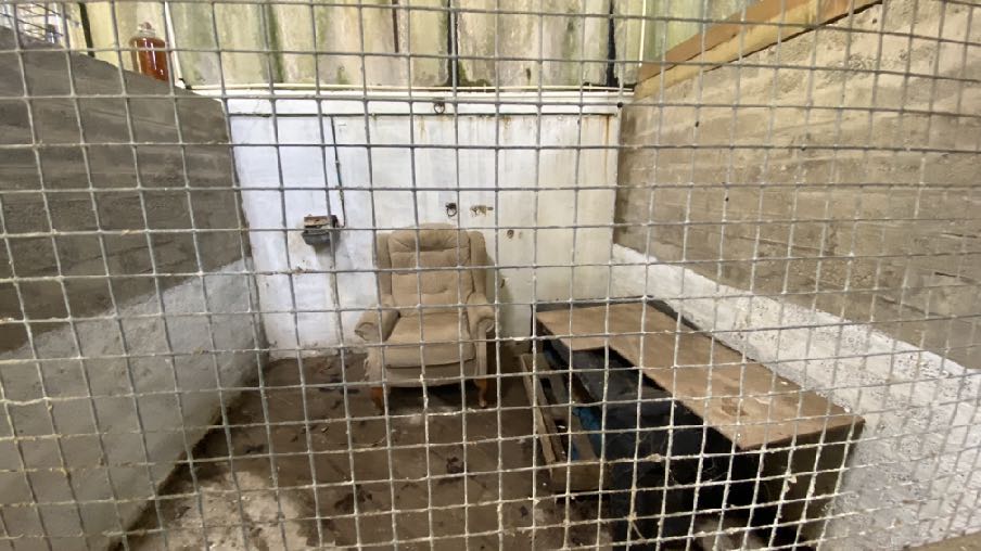 A photograph showing the interior of a derelict concrete block outbuilding viewed through a wire mesh fence, containing an armchair and a wooden table.