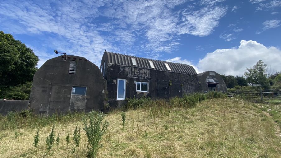 A photograph showing a large, dark concrete agricultural building with a curved roof, likely a Nissen hut, situated in a grassy field with new windows installed.