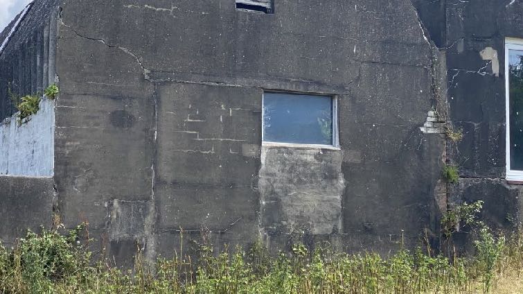 A close-up photograph of the weathered dark exterior wall of an existing outbuilding, featuring a window and vegetation growing at the base.