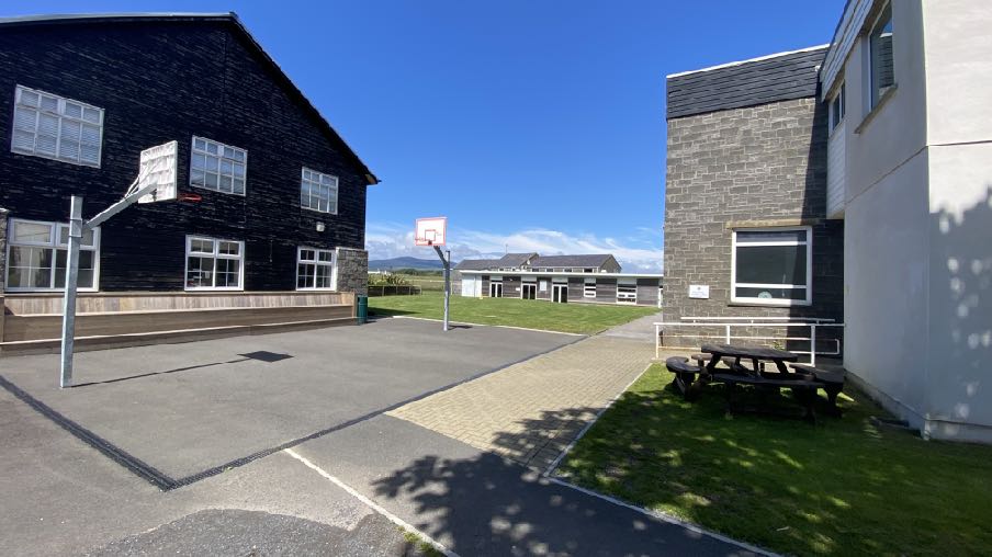 A photograph showing an outdoor paved playground area with basketball hoops situated between two buildings, featuring grass landscaping and a picnic table.