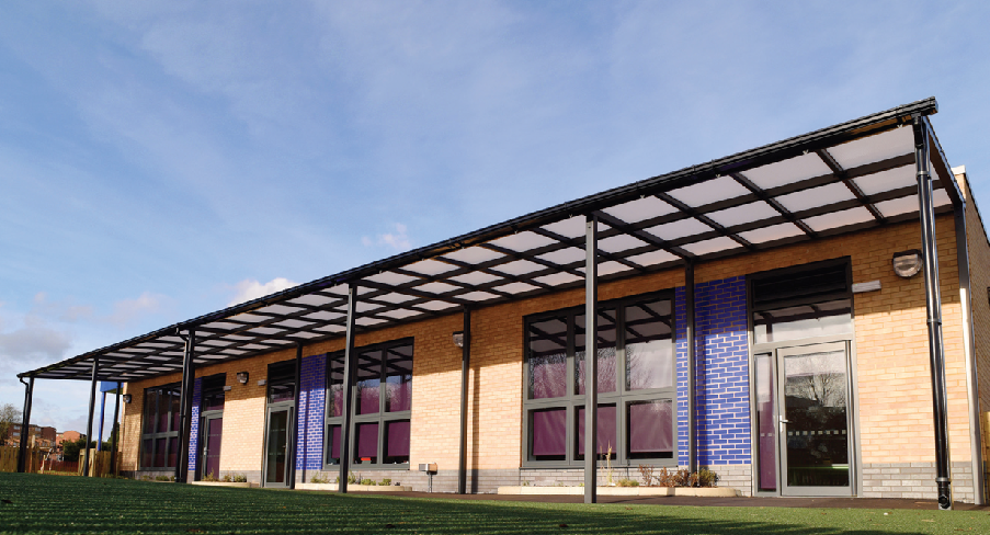 A photograph showing the exterior of a modern brick building, likely a school, featuring a large steel canopy structure and green landscaping in the foreground.