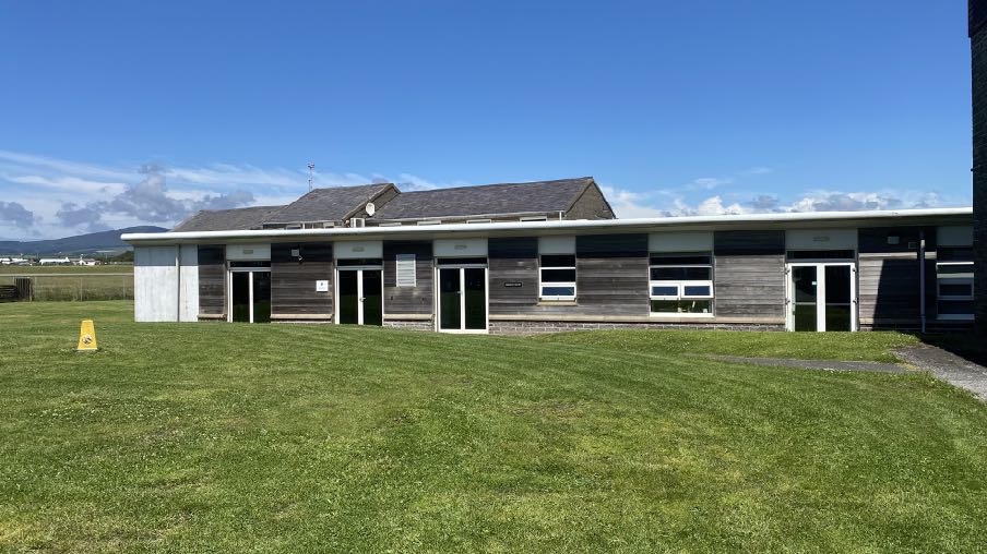 A photograph showing the exterior of a single-story building with wooden cladding and a flat canopy structure running along the front elevation, set against a grassy foreground.