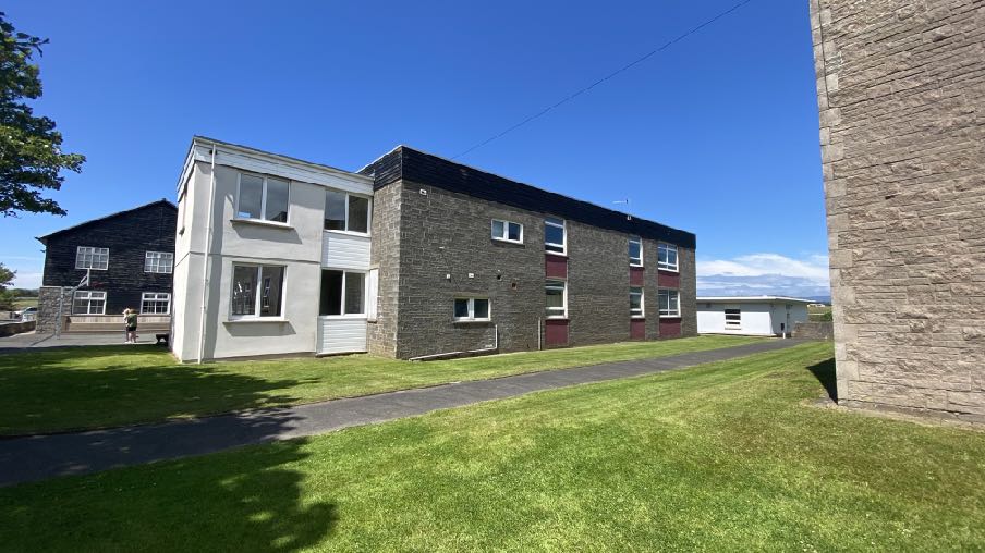 A photograph showing the exterior of a two-story building complex with mixed stone and rendered facades, set against a blue sky with a paved path and lawn in the foreground.