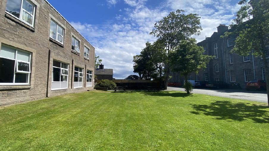 A photograph showing a grassy courtyard area between two stone buildings, likely part of a school site with cars parked in the background.