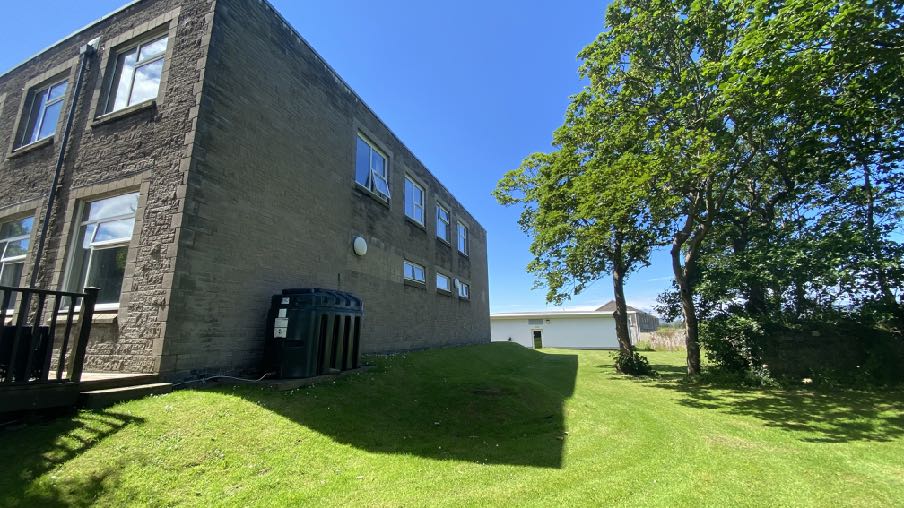 A low-angle outdoor photograph showing the exterior of a large stone building, likely Jackson House, with a grassy slope and trees in the foreground.
