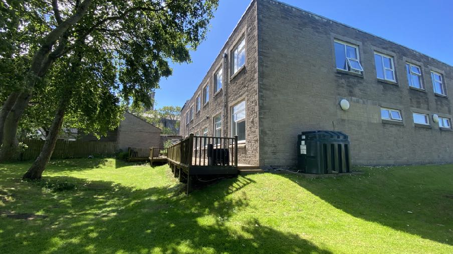 A photograph showing the exterior side elevation of a large stone building with a wooden deck and grassy grounds.