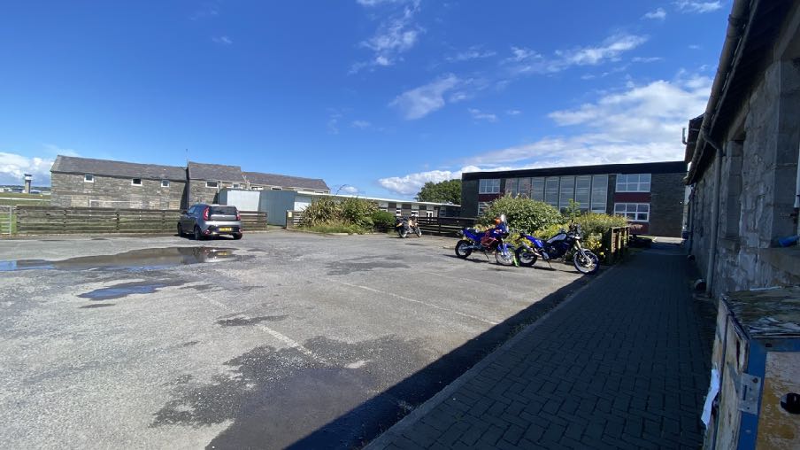 A photograph showing a paved yard or parking area with parked motorcycles and a car, bordered by stone buildings and a wooden fence under a blue sky.