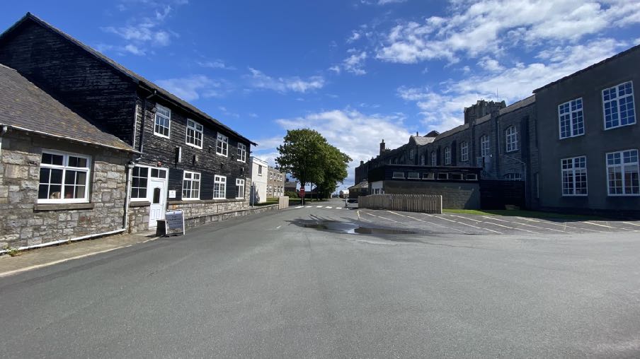 A street-level photograph showing stone buildings in Castletown, including Jackson House on the left and a larger institutional building on the right, with a paved parking area and wooden fencing.