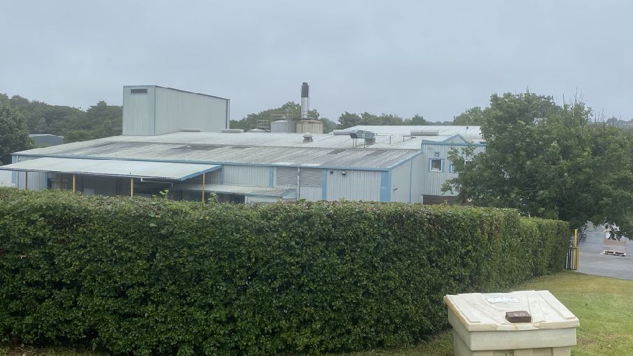 A photograph showing a large industrial or commercial building with a corrugated roof, viewed from behind a tall green hedge on an overcast day.