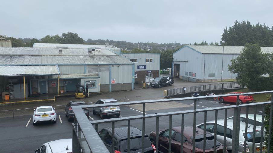 An elevated photograph showing a commercial or industrial estate with warehouse-style buildings, a parking lot with several cars, and a metal railing in the foreground.
