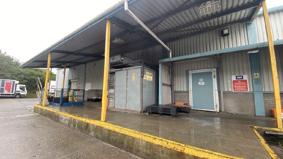 A photograph showing the exterior of a commercial or industrial building with a covered loading bay area. A large grey metal unit, likely a condenser, is installed under the canopy next to a blue door.