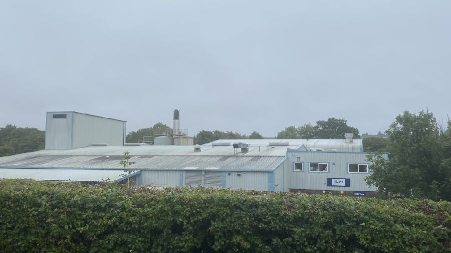A photograph showing a large industrial or commercial building with corrugated roofing and metal cladding, partially obscured by a green hedge in the foreground.
