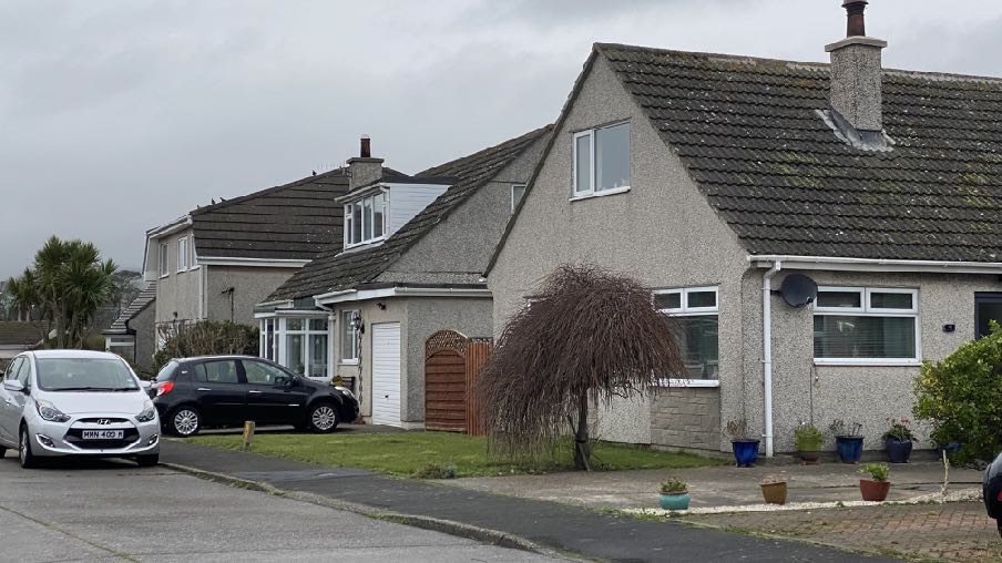 A street-level photograph showing a residential bungalow with a garage and driveway, alongside neighboring houses in a suburban setting.