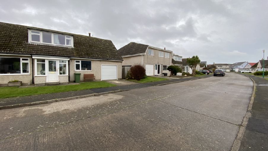 A street-level photograph showing a detached bungalow with a garage and driveway, situated on a residential road with other houses in the background.