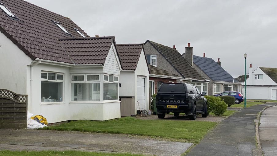 A street-level photograph showing a white residential property with a prominent front extension and an attached garage, with a black pickup truck parked in the driveway.