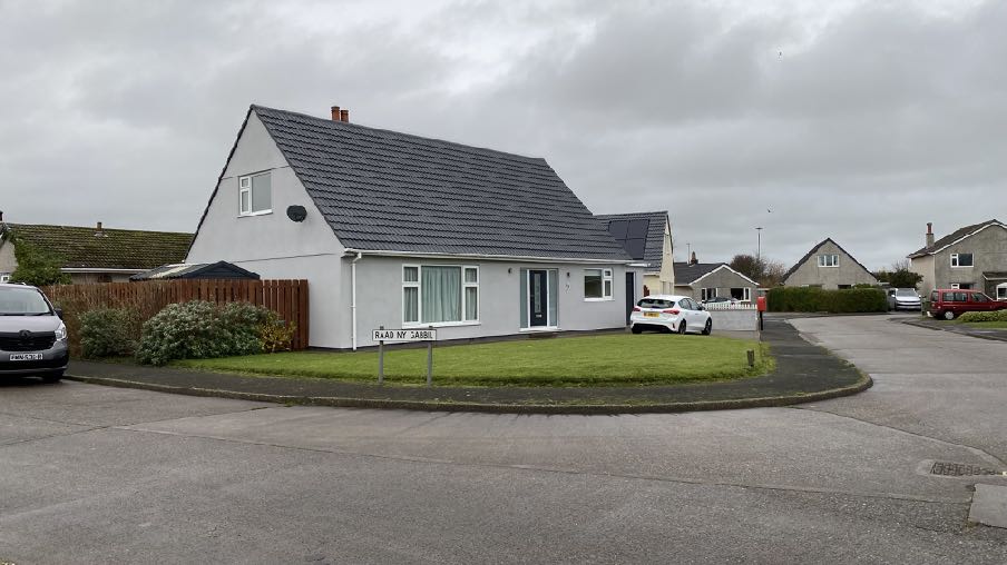 A street-level photograph of a detached white bungalow with a grey tiled roof. The property features a driveway with a parked car and a wooden boundary fence.