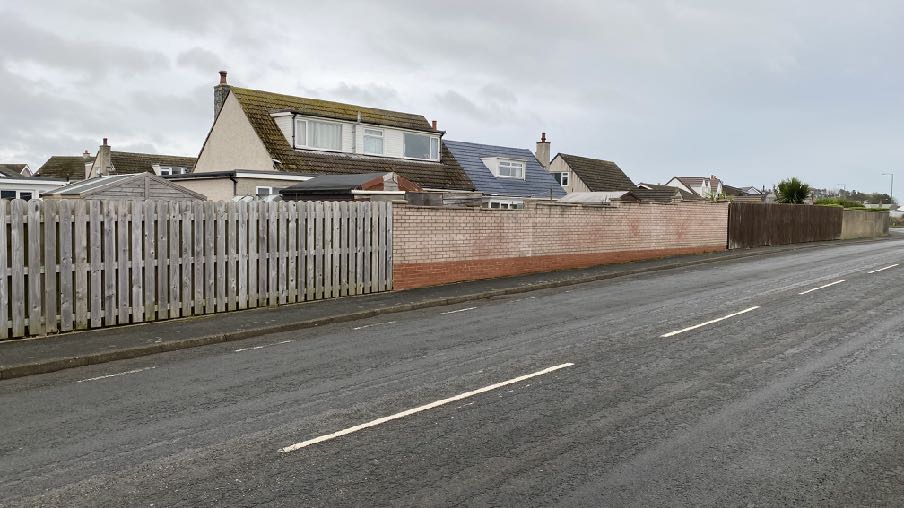 A street-level photograph showing a residential property boundary with a wooden fence and brick wall, with houses visible behind.