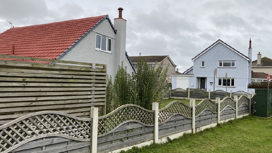 A street-level photograph showing a residential property with a red tiled roof behind a wooden fence, with neighboring houses visible in the background.