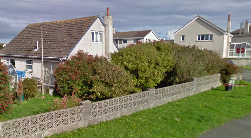 A street-level photograph showing a residential property with a tiled roof and chimney, partially obscured by a large hedge and a decorative concrete block boundary wall.