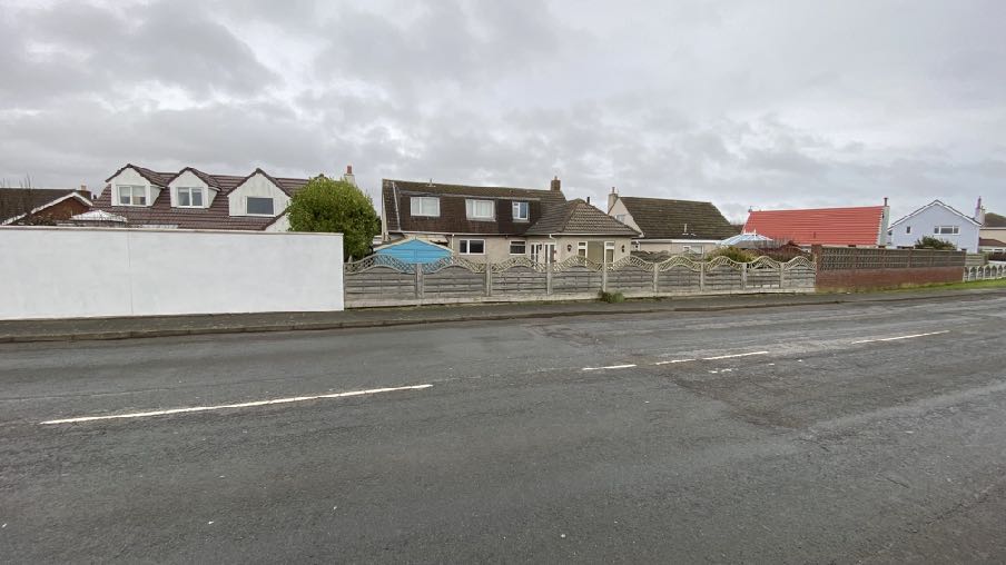 A street-level photograph showing a row of residential houses behind a white wall and fence under an overcast sky.