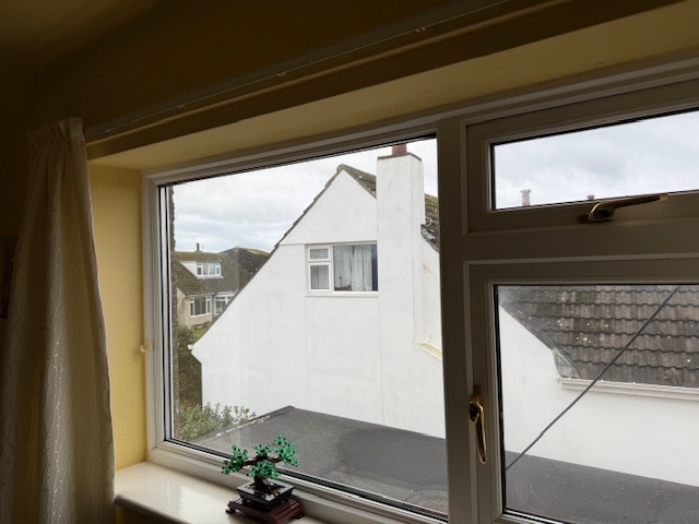 A photograph taken from inside a room looking out through a white window frame at a neighboring white house with a pitched roof.