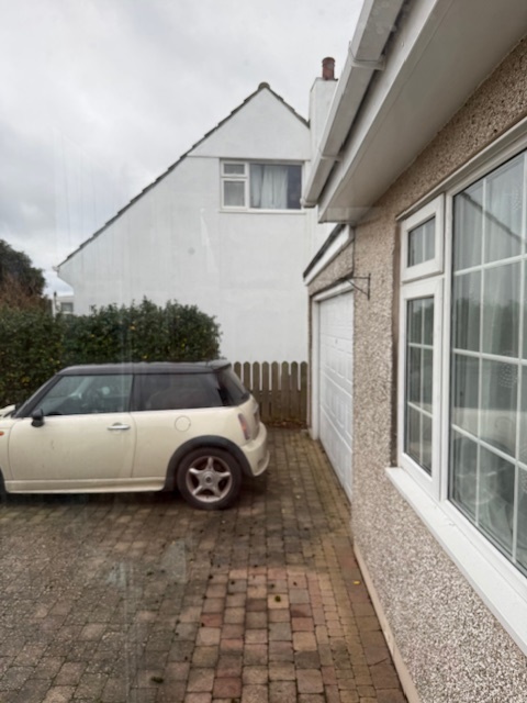 A photograph showing the existing side elevation of a residential property with a paved driveway, a white car, and a garage.