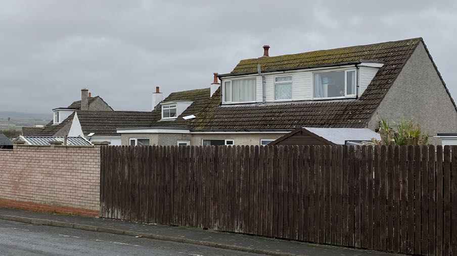 A street-level photograph showing a residential house with a large dormer window, viewed from behind a wooden fence and brick wall.