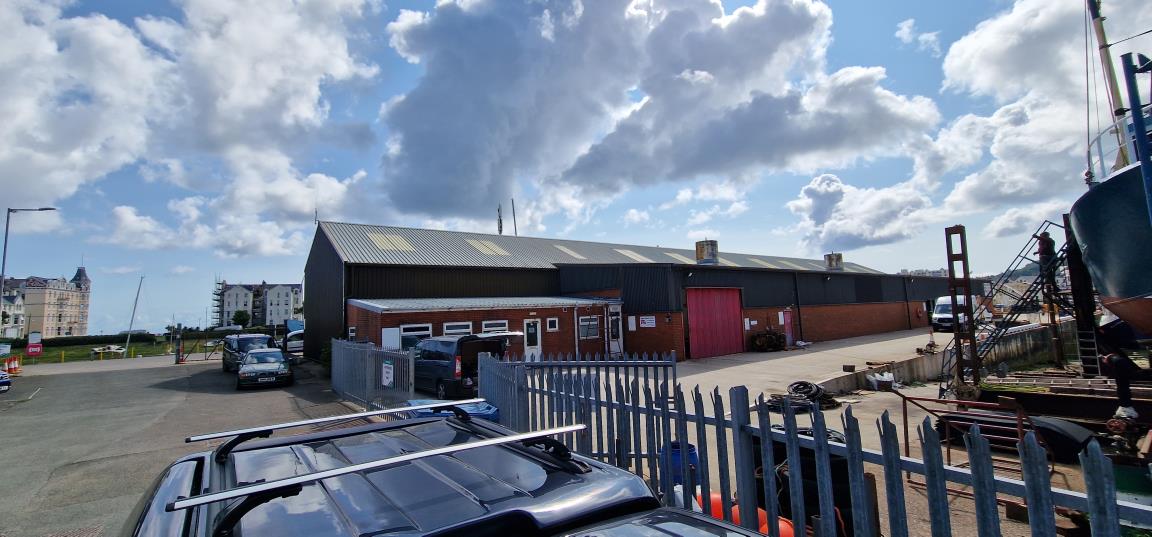 A photograph of an industrial site featuring a large dark building with a brick extension, a metal fence in the foreground, and a boat hull visible on the right.