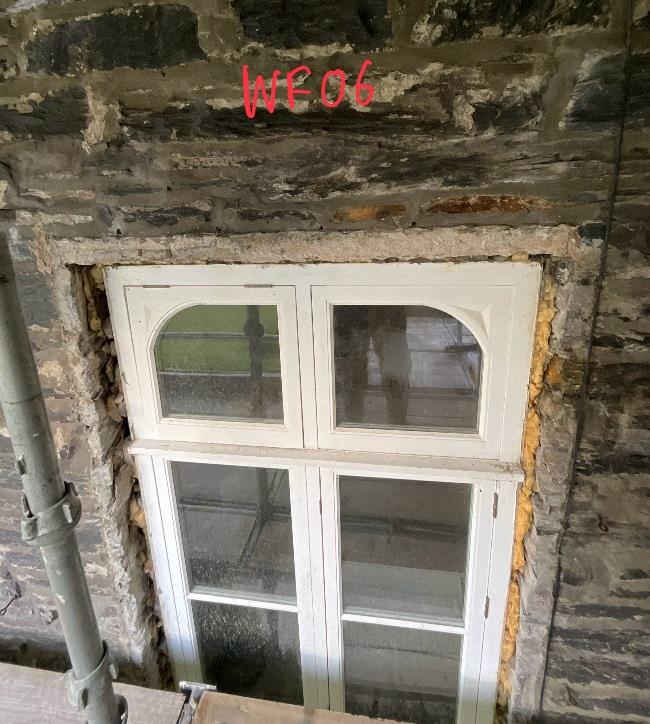 A close-up photograph of a white sash window set into a rough stone wall, showing scaffolding and yellow insulation foam around the frame.