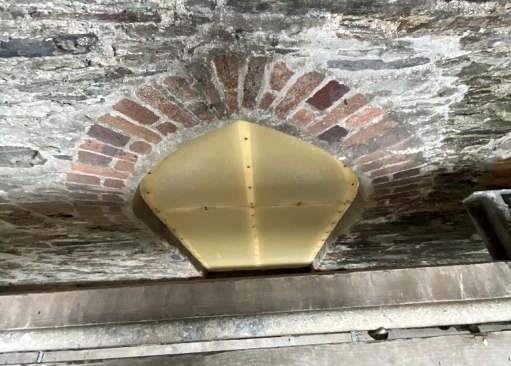 A close-up photograph of a weathered brick wall featuring a relieving arch and a brass metal cover plate.