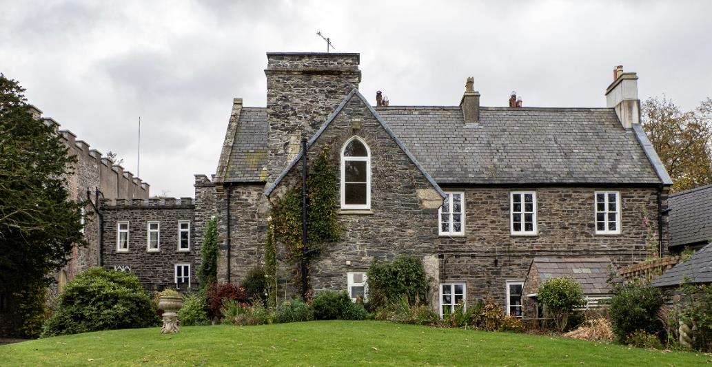 A photograph showing the exterior of a large stone building, likely a converted historic structure, featuring a prominent chimney stack on the gable end and slate roofing.