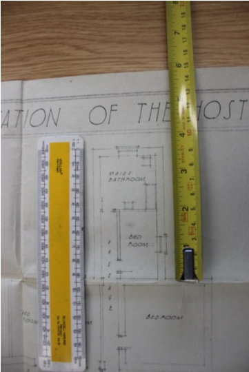 A photograph of an architectural floor plan showing room layouts like 'Main Bathroom' and 'Bed Room', with rulers and a tape measure placed on top for scale.