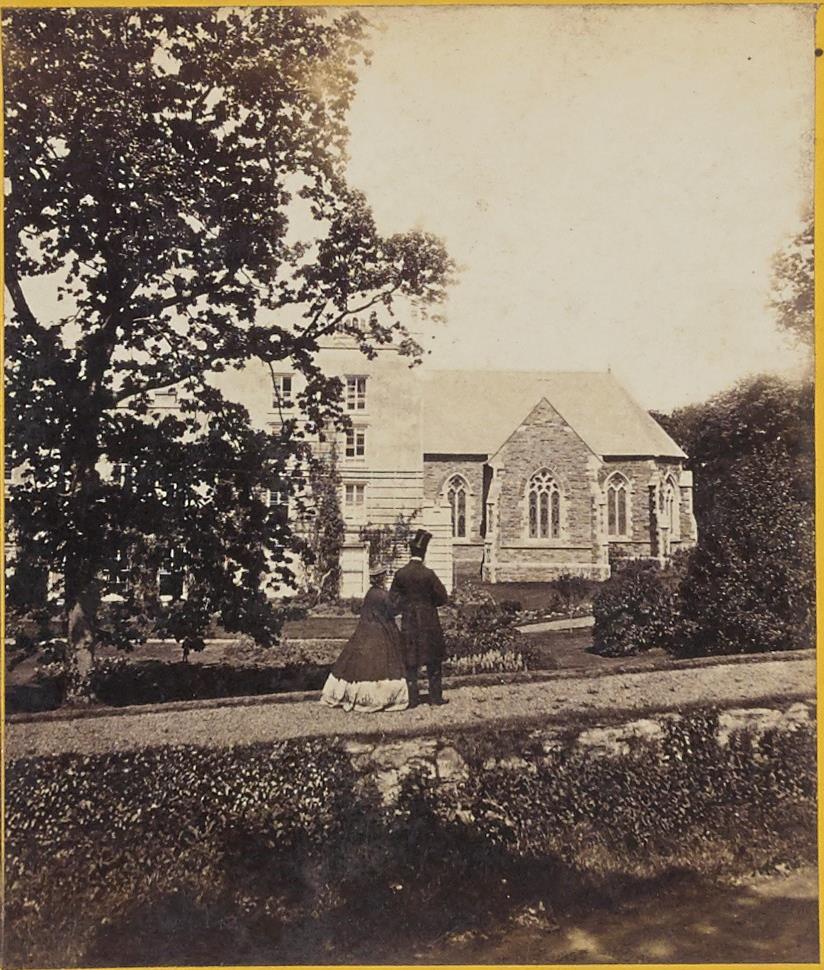 A vintage sepia-toned photograph showing a large stone building complex featuring a chapel-like structure with Gothic windows, viewed from a garden path with two Victorian-era figures in the foreground.