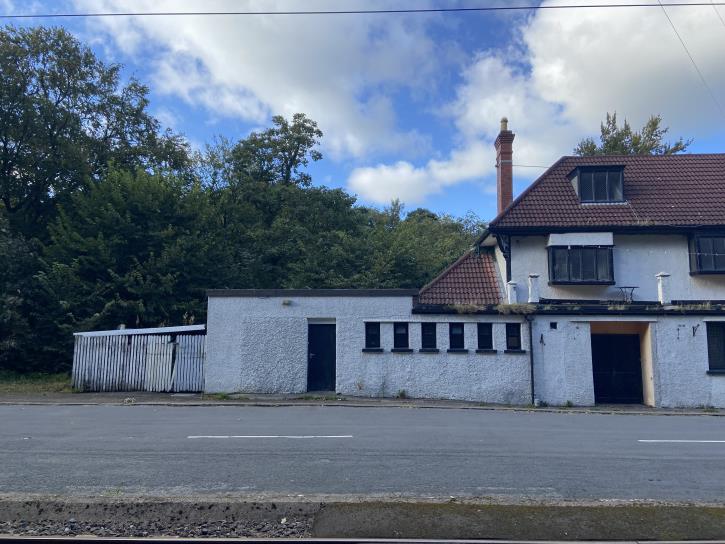 A street-level photograph showing a white single-storey flat-roofed structure attached to a larger two-storey building with a tiled roof, set against a backdrop of trees.