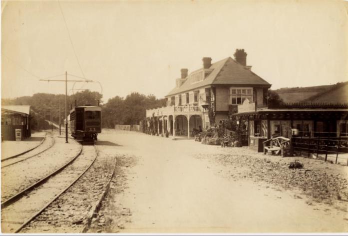 A vintage sepia-toned photograph depicting a street scene with tram tracks on the left and a large building with a balcony on the right.