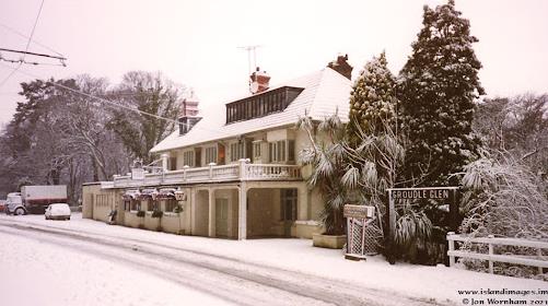 A snowy photograph of a large detached building with a balcony and pitched roof, situated next to a road with a sign reading 'SNOODLE GLEN'.