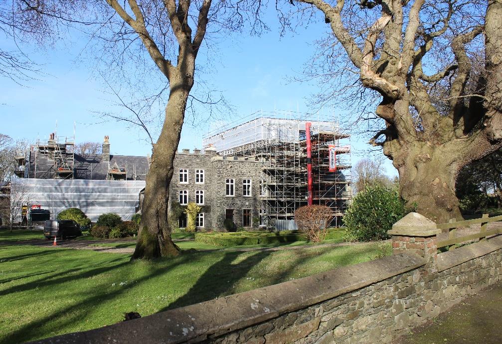 A photograph showing a large stone building undergoing significant renovation, covered in scaffolding, set within a rural landscape with trees and a stone wall.