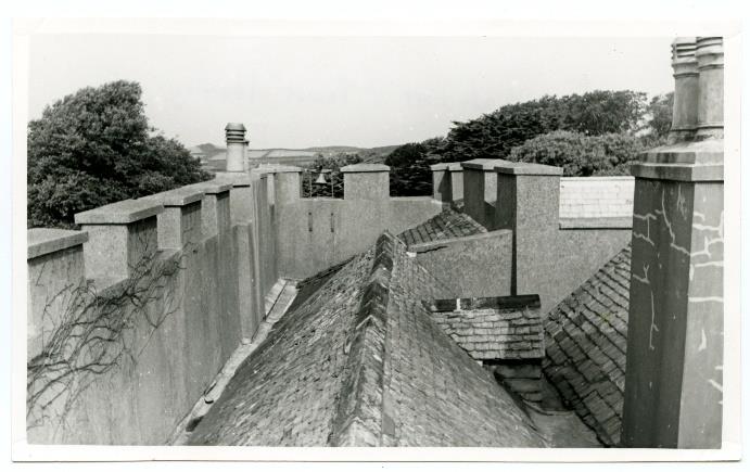 A black and white photograph showing a high-angle view of tiled roofs and multiple stone chimney stacks, including a prominent stack on the right and a long wall or parapet on the left.