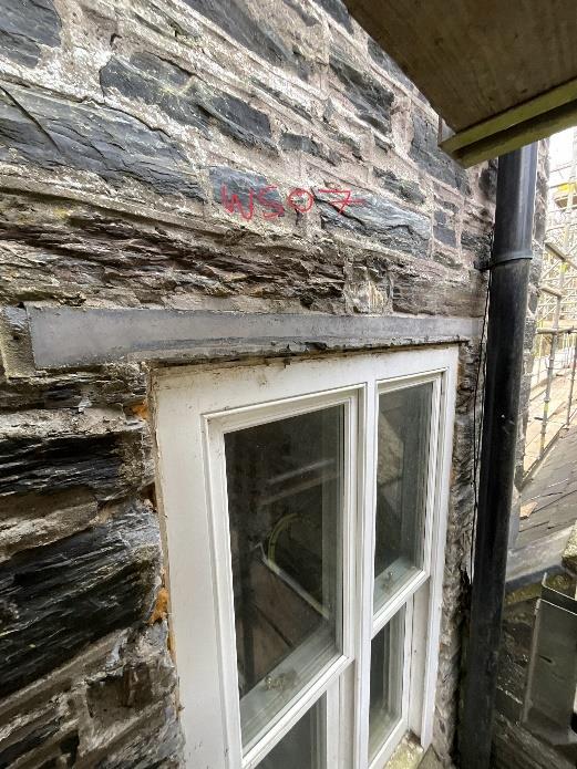 A close-up photograph of a historic slate stone wall featuring a white sash window, a grey lintel with red spray paint markings, and a black downpipe, with scaffolding visible in the background.