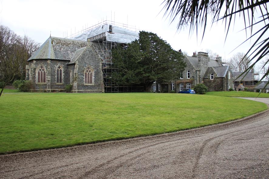 A photograph of a large stone building complex featuring Gothic-style windows and scaffolding on the roof, set on a green lawn with a gravel driveway.