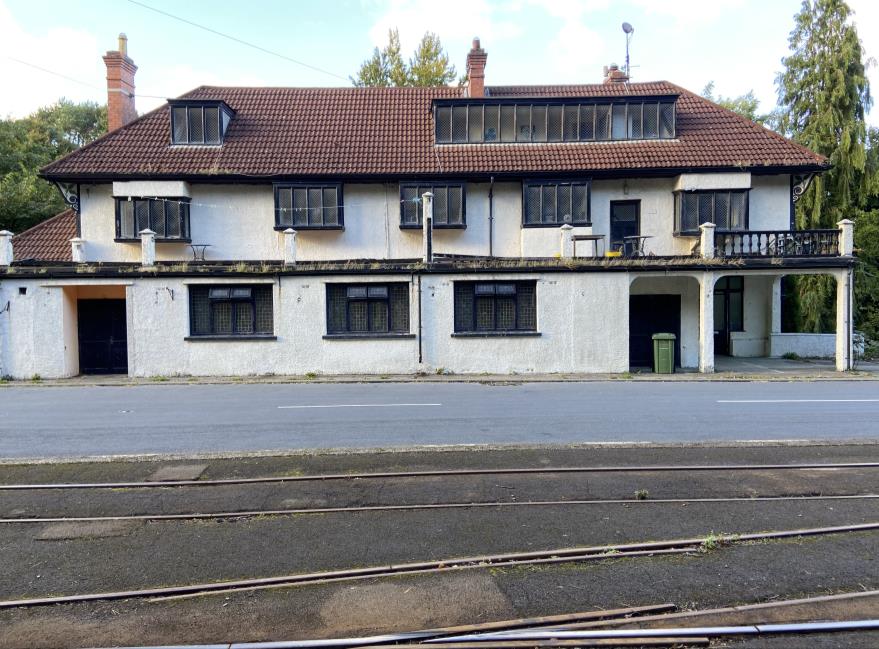 A photograph showing a large white two-story building with a brown tiled roof, situated behind tram tracks.