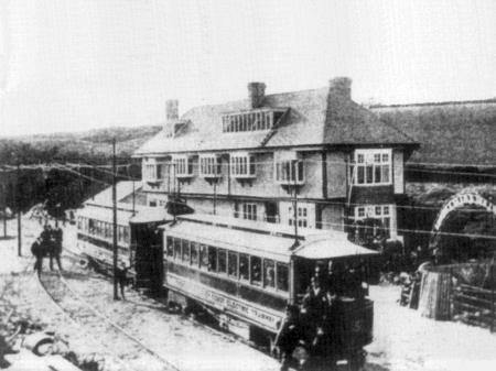 A grainy black and white historical photograph showing a large two-story building adjacent to a tram on tracks with people standing nearby.